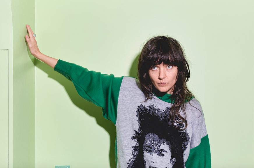 Die Sängerin Courtney Barnett lehnt sich vor grünem Hintergund an eine Wand an und trägt einen grün-grauen Pulli mit Aufdruck.