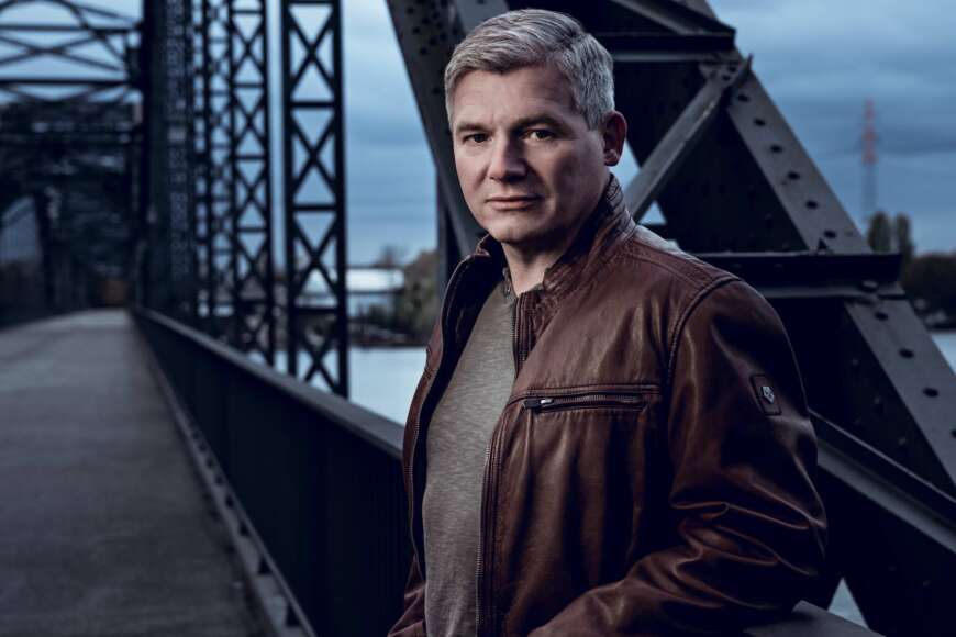 Autor Andreas Winkelmann ist mit grauen Haaren in einer braunen Lederjacke auf einer Stahlbrücke zu sehen.