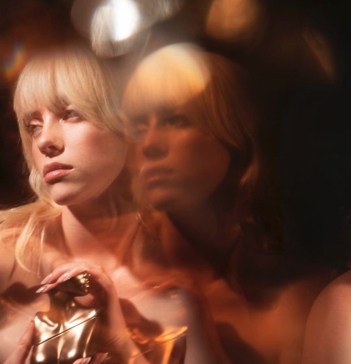 „TV“: Man sieht Billie Eilish mit blonden Haaren und einem torsoförmigen Perfumflakon in der Hand, den sie selbst entworfen hat.