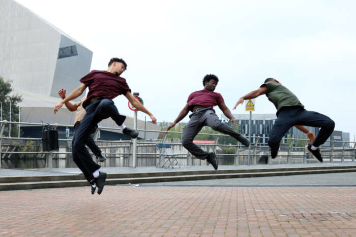Just us Dance Theatre: Vier junge Männer tanzen auf der Straße, sie sind synchron in einer gesprungenen Drehung.