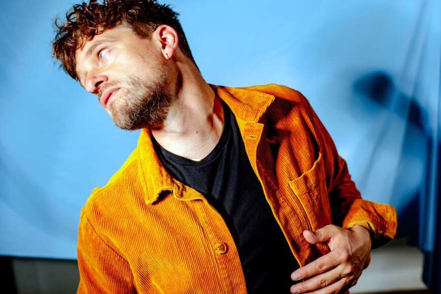 Zu sehen ist der dänische Künstler Lasse Matthiessen, der für seine neue Single "Emotional" in orangefarbenem Cord-Hemd vor blauem Himmel posiert.