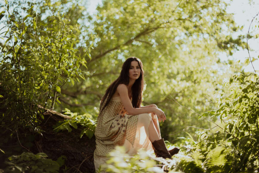 Lena Meyer-Landrut in einem langen, sommerlichen Kleid, sie sitzt auf einem Baumstamm umgeben von Natur und grün und schaut in die Kamera.