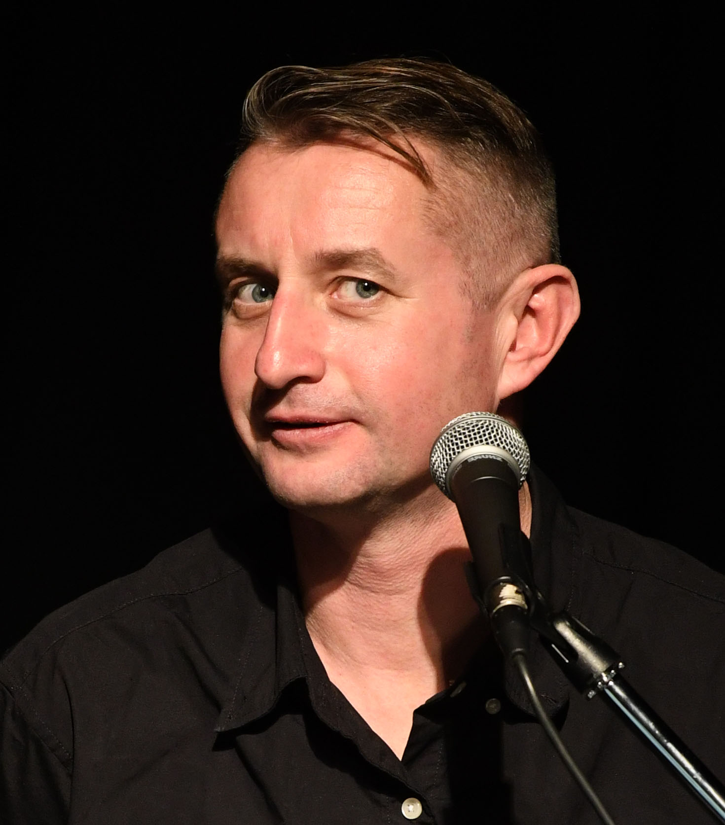 Zu sehen ist der Autor Serhiy Zhadan in schwarzem hemd vor schwarzem Hintergrund, er guckt vor einem Mikrofon stehend verschmitzt in die Kamera.