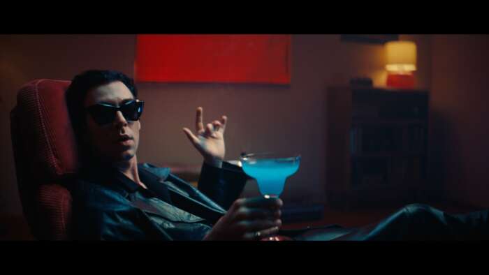 Ein Still aus dem Video „Space up“ von Niqtin ist zu sehen, in dem der Sänger mit Sonnebrille auf einem Sessel sitzt und einen knallblauen Drink aus einem Stielglas trinkt.