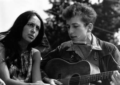 Ein schwarz-weißes Foto von Joan Baez und Bob Dylan. Dylan hat eine Gitarre im Arm und eine Mundharmonika vor dem Mund.
