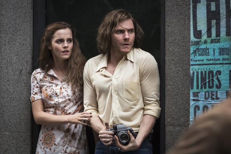 Still aus „Colonia Dignidad“: Emma Watson und Daniel Brühl stehen nebeneinander in einem Hauseingang, sie wirken panisch. Watson hält sich an Brühls Arm fest, der eine Kamera in den Händen hält.