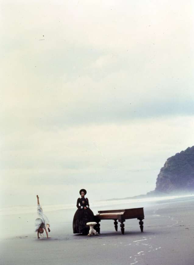 Still aus dem Film „Das Piano“: An einem diesigen Strand von Neuseeland sitz eine Frau am Klavier und ein Kind macht einen Handstand.