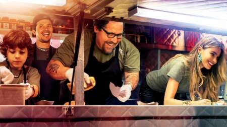 Still aus „Kiss the Cook“: Percy (Emjay Anthony, l.), Martin (John Leguizamo, 2.v.l.), Carl (Jon Favreau, 2.v.r.) und Carls Ex-Frau Inez (Sofía Vergara)lehnen sich lächelnd aus einem Food Truck.