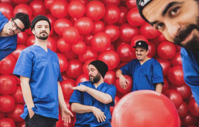 Pressefoto Le Fly für L'Amour, Album und Tour: Zu sehen sind fünf Bandmitglieder in knallblauen Shirts, die vor roten Luftballons Grimassen ziehen.