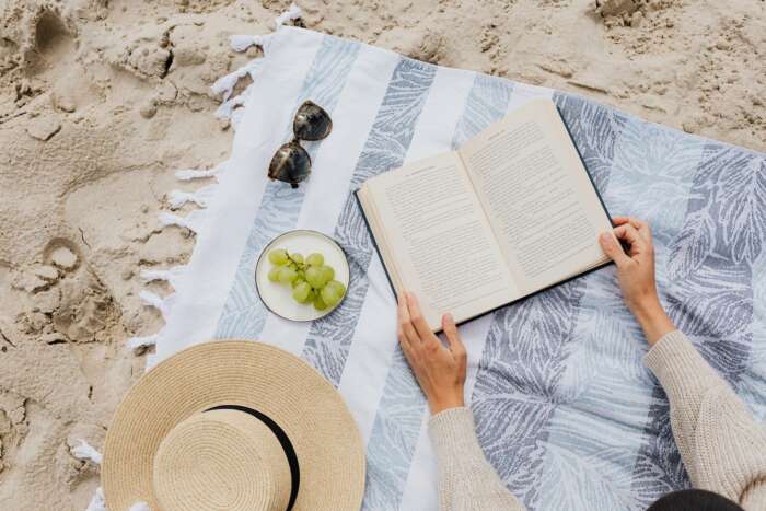 Bücher für den Sommer 2022: Zwei Hände schlagen ein Buch auf einer Picknickdecke auf, die am Sandstrand liegt.