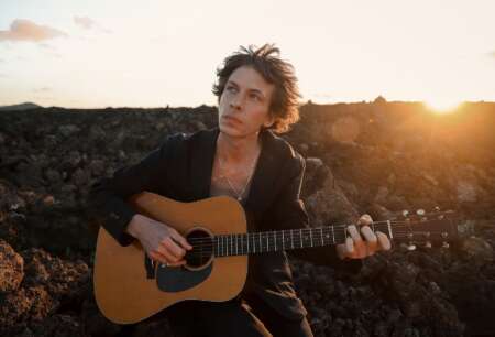 Zu sehen ist Musiker Max Prosa mit Akustikgitarre vor einem goldglänzenden Sonnenuntergang.