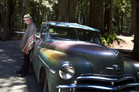 Neil Young ist zu sehen, wie er, mit einem bunten Poncho bekleidet, an einem schillernden Oldtimer lehnt, der in einem Wald zwischen Bäumen geparkt ist.