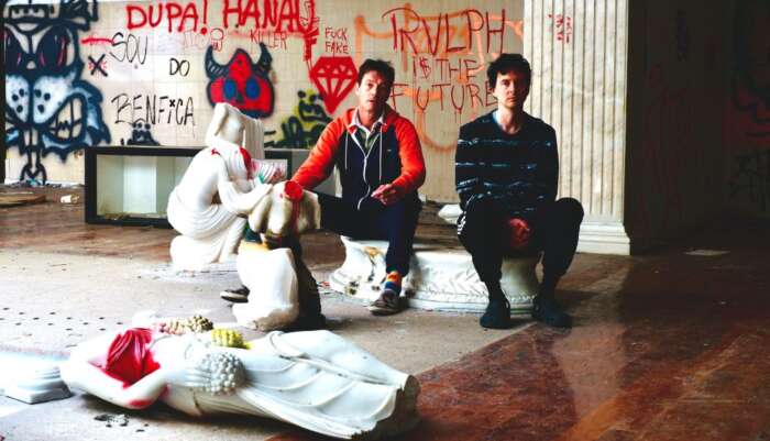 Panda Bear und Sonic Boom sitzen auf einem Sockel vor einer Wand mit Graffiti. Um sie herum liegen zerstörte antike Statuen.