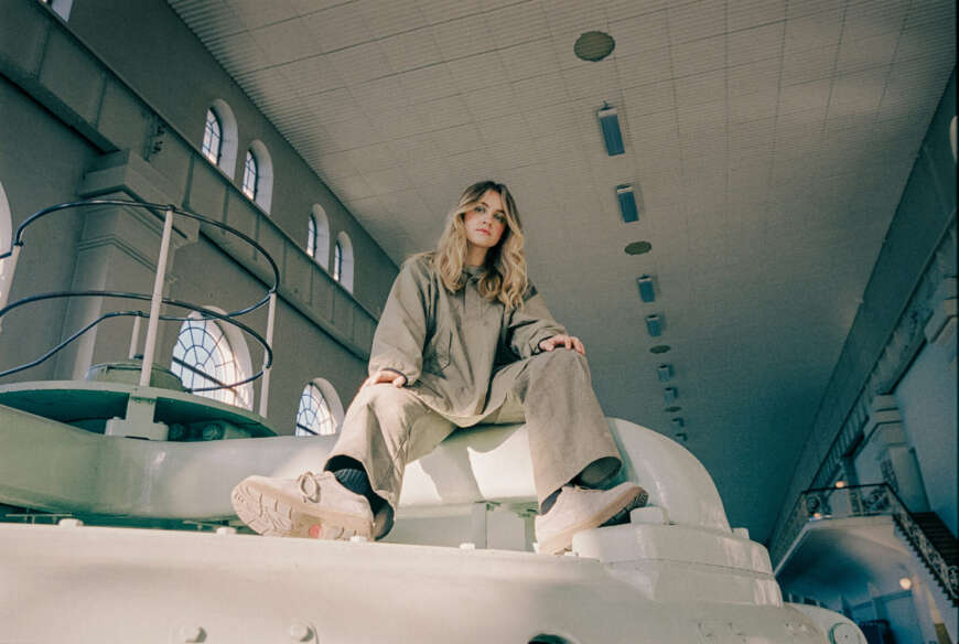 Zu sehen ist die Sängerin Skaar in einem beigen Outfit, dicken, weißen Sneakers. Sie sitzt auf einer Art weißen Podest und schaut von oben in die Kamera.
