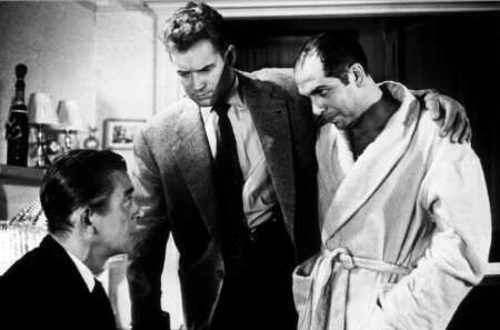 Still aus „Rififi“: Tony sitzt auf einem Stuhl und blickt zweifelnd zu Jo und Mario, die über ihm stehen.