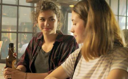 Still aus „Siebzehn“: Zwei Teenagerinnen sitzen nebeneinander an einer Theke. Die linke hält eine Bierflasche in der Hand und lächelt die rechte an.