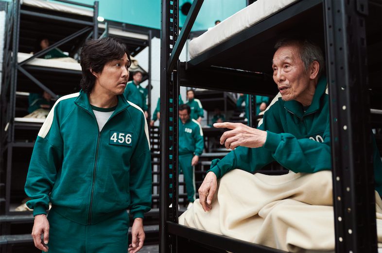 Still aus „Squid Game“: Lee Jung-jae steht neben Oh Young-soos Hochbett. Sie beide tragen grüne Overalls mit Nummern auf der Brust.