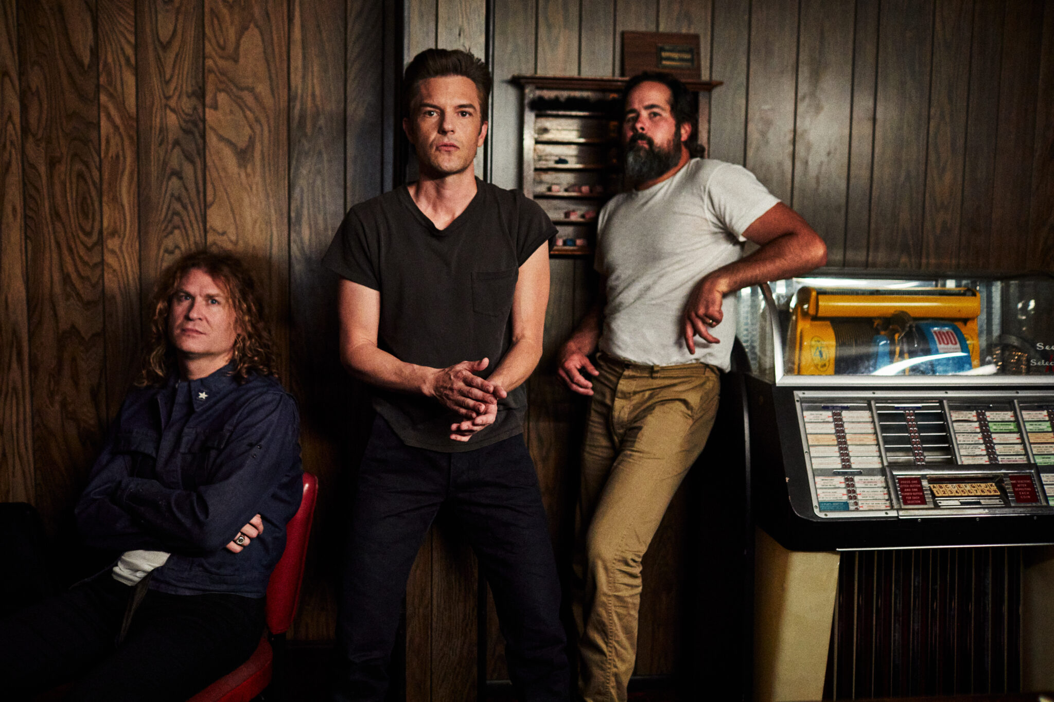 „The Killers“: Man sieht von links Dave Keuning, Brandon Flowers und Ronnie Vannucci Jr. vor einer dunklen Holzwand, Keuning sitzt, Flowers steht gegen die Wand gelehnt und Vannucci lehnt gegen eine Jukebox am rechten Bildrand.