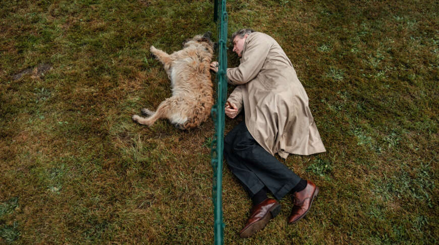 „Ruhe! Hier stirbt Lothar“: Man sieht Lothar (Jens Harzer) auf der einen Seite vom Zaun liegend und sein ehemaliger Hund auf der anderen Seite liegend (Draufsicht).