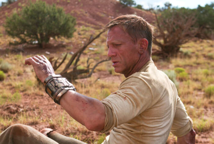 „Cowboys & Aliens“ auf Kabel eins: Man sieht Daniel Craig, der ein technischen Armreif an deinem Arm betrachtet, er ist in der Wüste, sitzt auf dem Boden und ist beschmutzt.