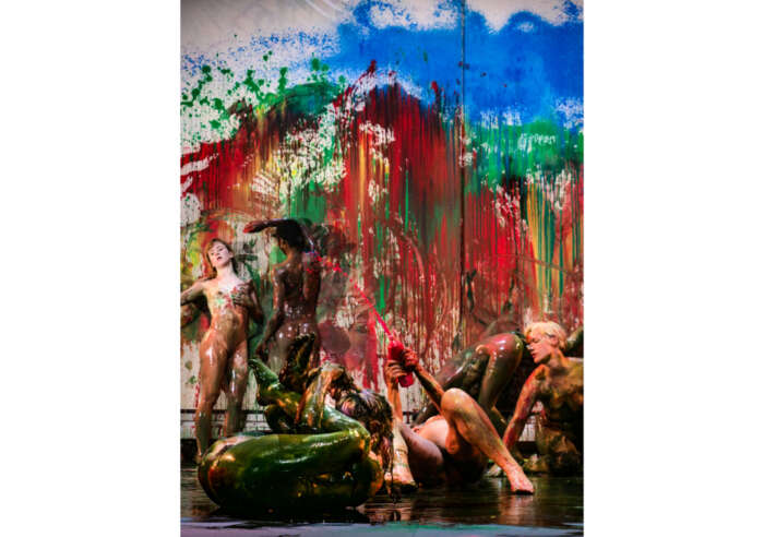 Eine Gruppe nackter mit bunter Farbe beschmierter Tänzerinnen und Tänzer auf einer Bühne