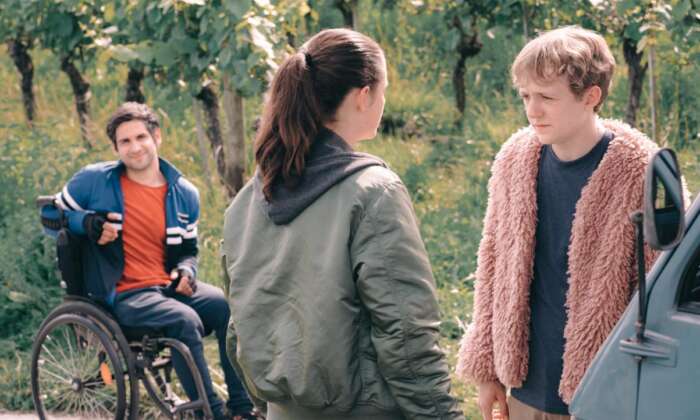 Still aus „Sommer auf drei Rädern“: Ein Mädchen und ein Junge stehen sich gegenüber. Ein dritter Junge im Rollstuhl lächelt ihnen zu.