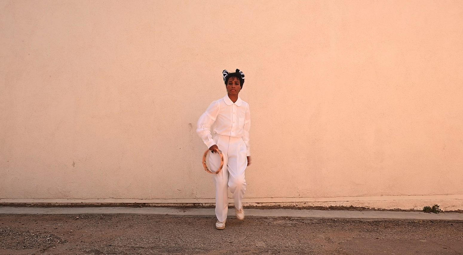 Santigold steht mit Schellenkranz in der Hand vor einer Wand