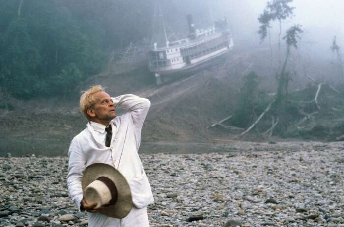 Klaus Kinski als Fitzcarraldo mit weißem Anzu und Hut in der Hand. Im Hintergrund liegt ein Dampfer zwischen Bäumen im Urwald.