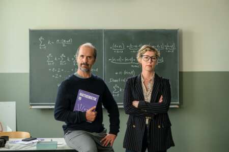 "Lehrer kann jeder!": Richard Glossat (Christoph Maria Herbst) sitzt in einem Klassenzimmer auf dem Lehrerpult und hält ein Mathematik-Buch in der Hand. Neben ihm steht Anke Glossat (Brigitte Zeh) mit verschränkten Armen und genervtem Blick. Im Hintergrund eine Schultafel, an die mit Kreide Mathematik-Formeln geschrieben stehen.