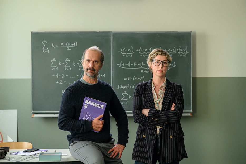 "Lehrer kann jeder!": Richard Glossat (Christoph Maria Herbst) sitzt in einem Klassenzimmer auf dem Lehrerpult und hält ein Mathematik-Buch in der Hand. Neben ihm steht Anke Glossat (Brigitte Zeh) mit verschränkten Armen und genervtem Blick. Im Hintergrund eine Schultafel, an die mit Kreide Mathematik-Formeln geschrieben stehen.