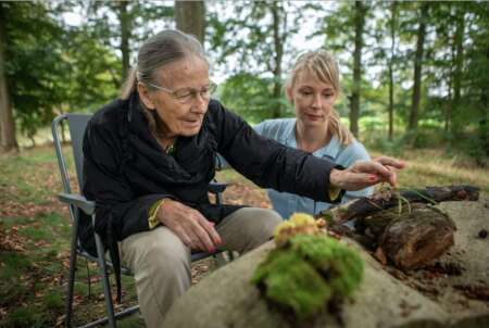 "Mitgefühl - Pflege neu denken": Alte Frau sitzt mit junger Frau vor einem großen Stein auf dem Gras.