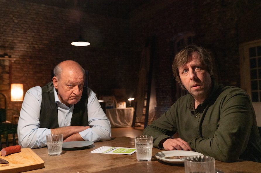 "Wilsberg – Schmeckt nach Mord": Wilsberg (Leonard Lansink) sitzt neben Ekki (Oliver Korittke) in seiner Halle an einem Holztisch und schaut kritisch geradeaus. Auf dem Tisch befindet sich ein Holzbrettchen, auf dem eine angeschnittene Wurst liegt.