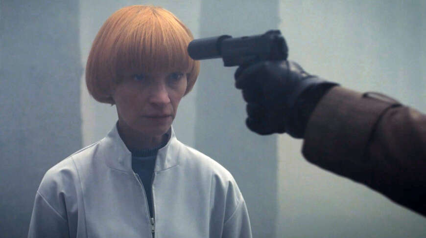 Die Wissenschaftlerin Edmunda (Agata Buzek) im weißen Kittel mit roten Haaren. Ihr wird eine Pistole an den Kopf gehalten.