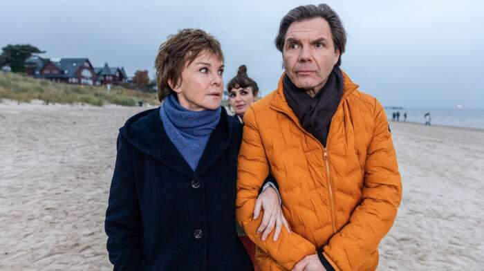 Still aus „Der Usedom-Krimi: Das Ende einer Reise“: Karin Lossow (Katrin Sass) und Dr. Brunner (Max Hopp) gehen Arm im Arm am Strand entlang.
