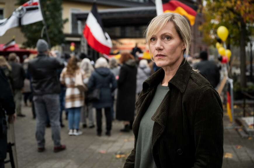 Still aus „Die Bürgermeisterin“: Claudia (Anna Schudt) blickt besorgt. Im Hintergrund ist eine Demonstration mit Deutschland- und Reichsflaggen zu sehen.