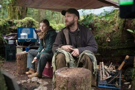 "Leave No Trace - Meine Wildnis": Tom (Thomasin McKenzie) und Will (Ben Foster) sitzen inmitten ihres selbst aufgebauten Waldlagers nebeneinander. Links neben Tom, die auf einer Plastikkiste sitzt, befindet sich ein Campingherd mit einem darauf stehenden Kochtopf. Zum Regenschutz hängt eine ausgespannte Plastikplane über den beiden. Rechts neben Will steht eine Kiste mit Feuerholz.