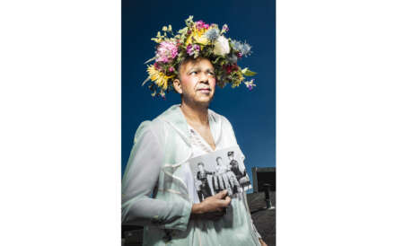 Ein Mann trägt ein Kleid, hat ein Foto in der Hand und einen Blumenkranz auf dem Kopf