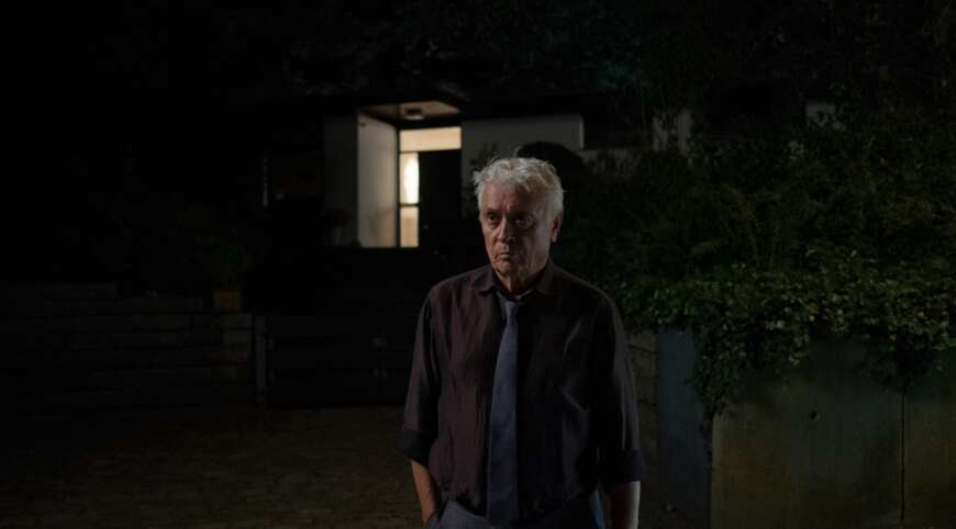 “Das Licht in einem dunklen Haus“: Johannes Fischer (Henry Hübchen) steht nachts vor einem Haus und blickt in die Ferne. Hinter ihm leuchtet aus der Eingangstür ein Licht.