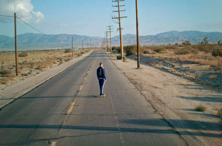 Still aus „Home“: Ein amerikanischer Highway in einer kargen Landschaft, darauf ist von hinten die Figur eines Mannes im Trainingsanzug zu sehen, der ein Skateboard fährt.