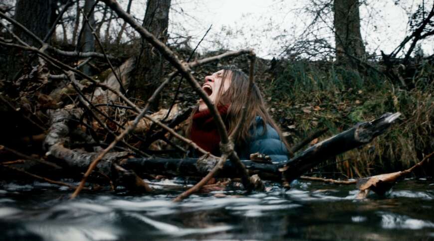 Still aus „Kalt“: Eine Frau steht in einem Fluss inmitten von Treibholz, ihr Gesicht ist verzweifelt verzerrt.