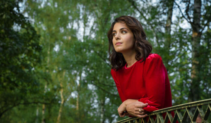 Katie Melua steht in einem Wald, hat ein rotes Shirt an und lehnt an einem Geländer.