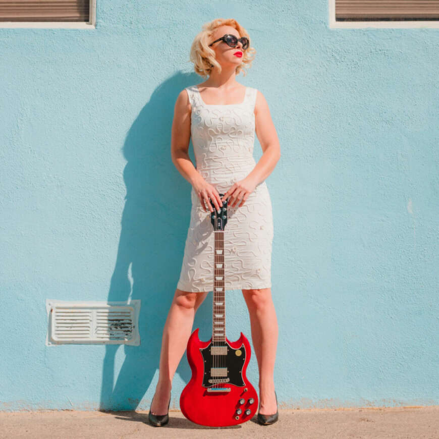 Samantha Fish steht mit einer roten Gitarre vor einer türkisfarbenen Wand.
