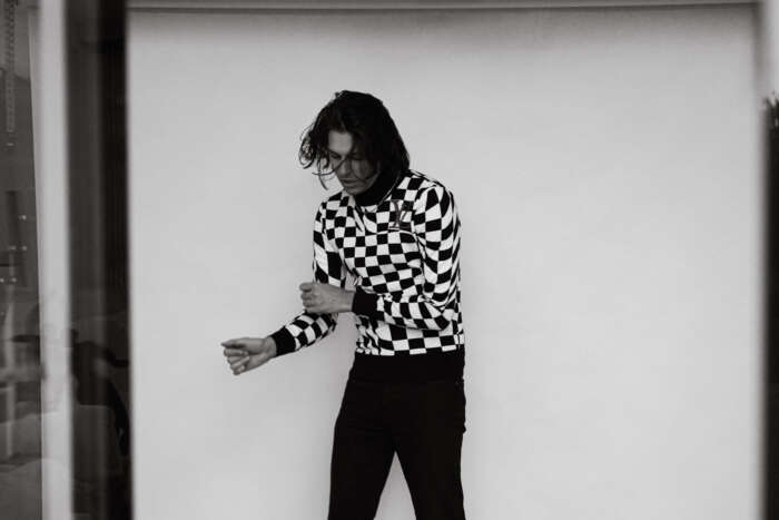 Ferdinand fka Left Boy steht vor einer Wand in einem schwarz-weiß karierten Sweatshirt.
