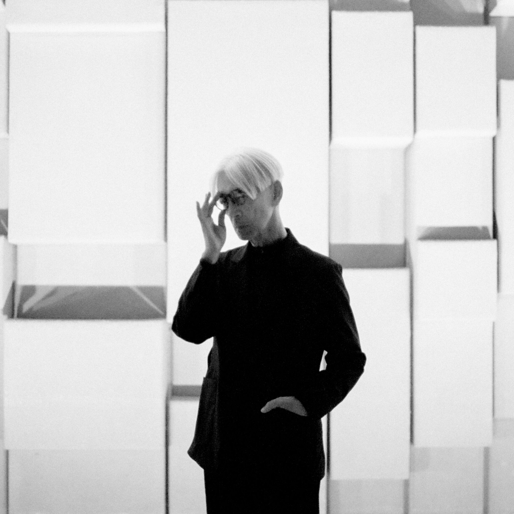 Ryuichi Sakamoto steht vor einer Wand und richtet seine Brille.