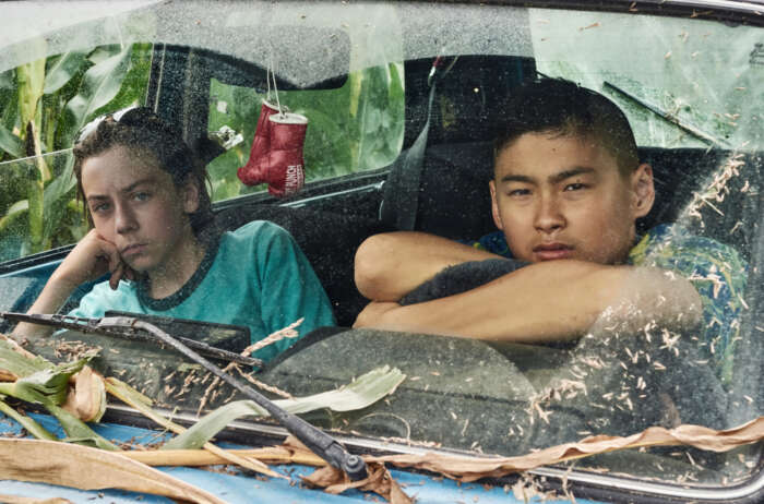 Still aus „Tschick“: Maik (Tristan Göbel) und Tschick (Anand Batbileg) sitzen hinter der Windschutzscheibe eines Autos. Die Kühlerhaube ist voller Blätter.