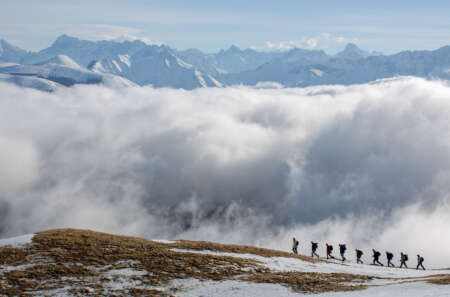 Pressebild „Auferstehen“: Eine kleine Reihe aus neun Männern geht einen Berghang hinauf. Dahinter sind eine riesige Wolkenbank und viele weitere Gipfel zu sehen.