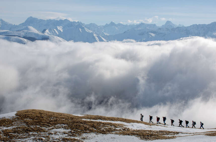 Pressebild „Auferstehen“: Eine kleine Reihe aus neun Männern geht einen Berghang hinauf. Dahinter sind eine riesige Wolkenbank und viele weitere Gipfel zu sehen.