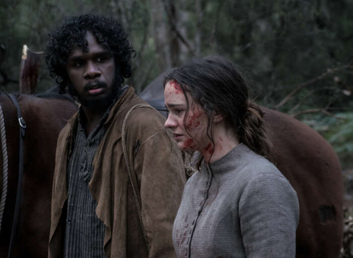 "The Nightingale": Clare (Aisling Franciosi) steht mit Blut verschmiertem Gesicht neben ihrem Begleiter, dem Aborigine Billy (Baykali Ganambarr), der sie entgeistert anschaut. Im Hintergrund ist der Rücken eines Pferdes im Wald zu sehen.