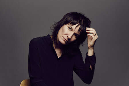 Jen Cloher sitzt auf einem Stuhl, hat eine Hand im Haar und schaut neutral in die Kamera.