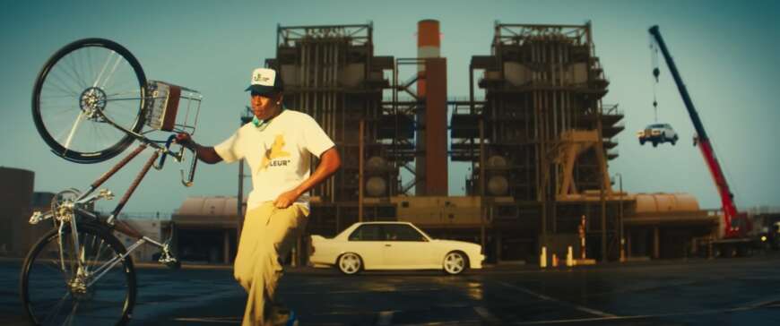 Tyler, The Creator steht vor einer Industrieanlage und neben einem Fahrrad.
